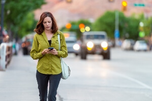 young woman looking down at her phone while walking near a busy street