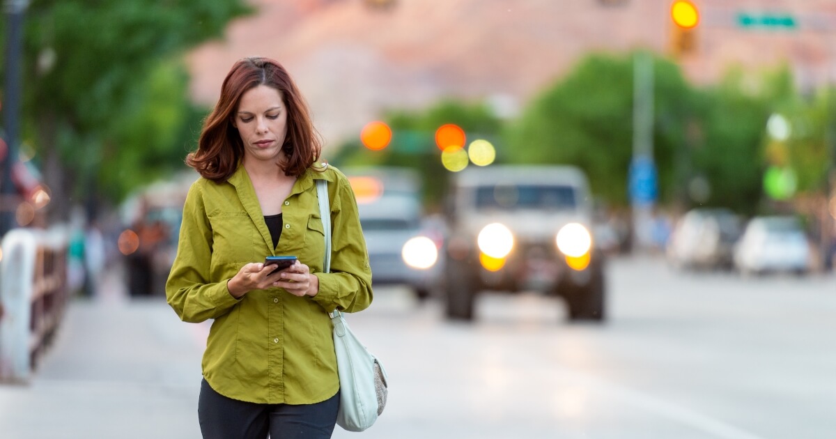 young woman looking down at her phone while walking near a busy street